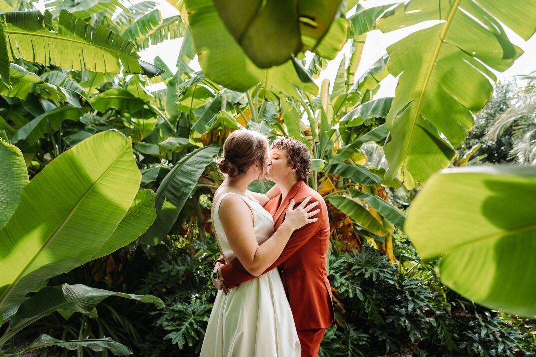 Two Brides, one in a white dress and one in an orange suit, kissing surrounded by Banana Tree Leaves